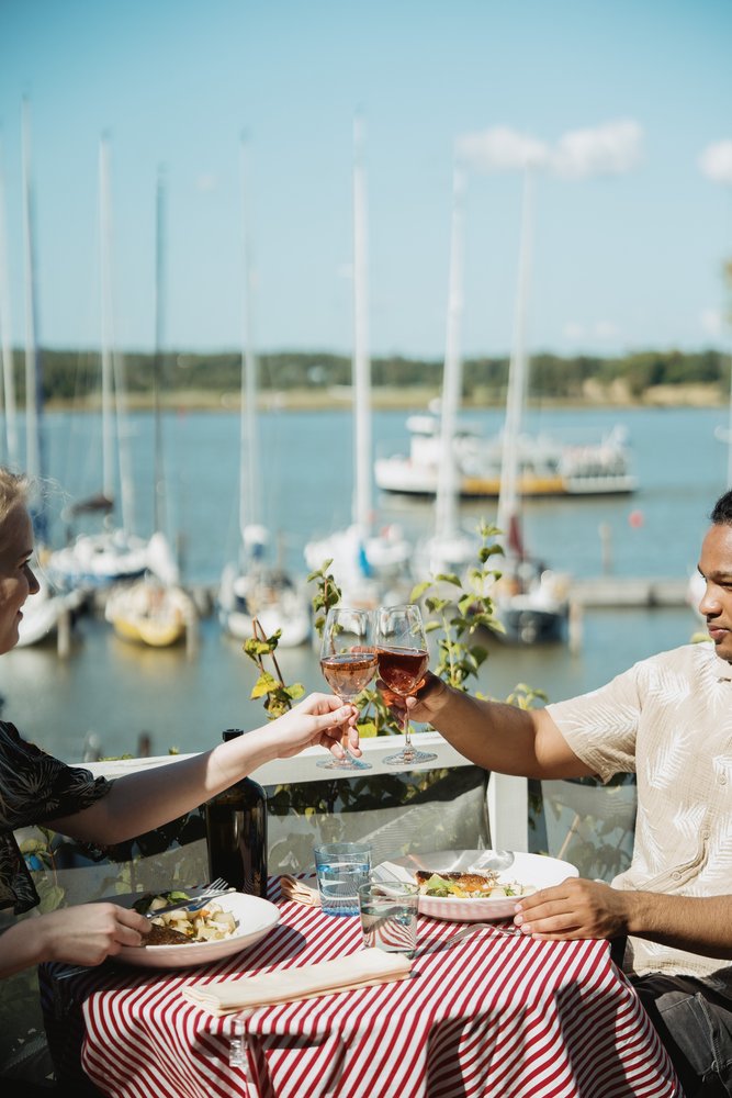 Two guests raising glasses at a harbour-side table overlooking moored yachts in the archipelago