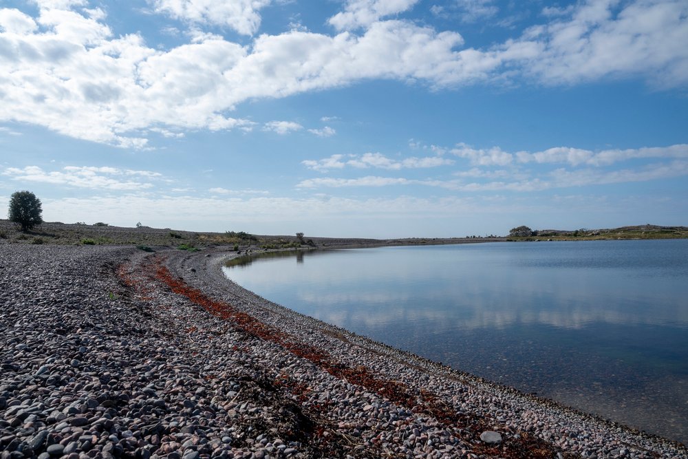 A wide pebble shore curving into still water under a high summer sky in the archipelago