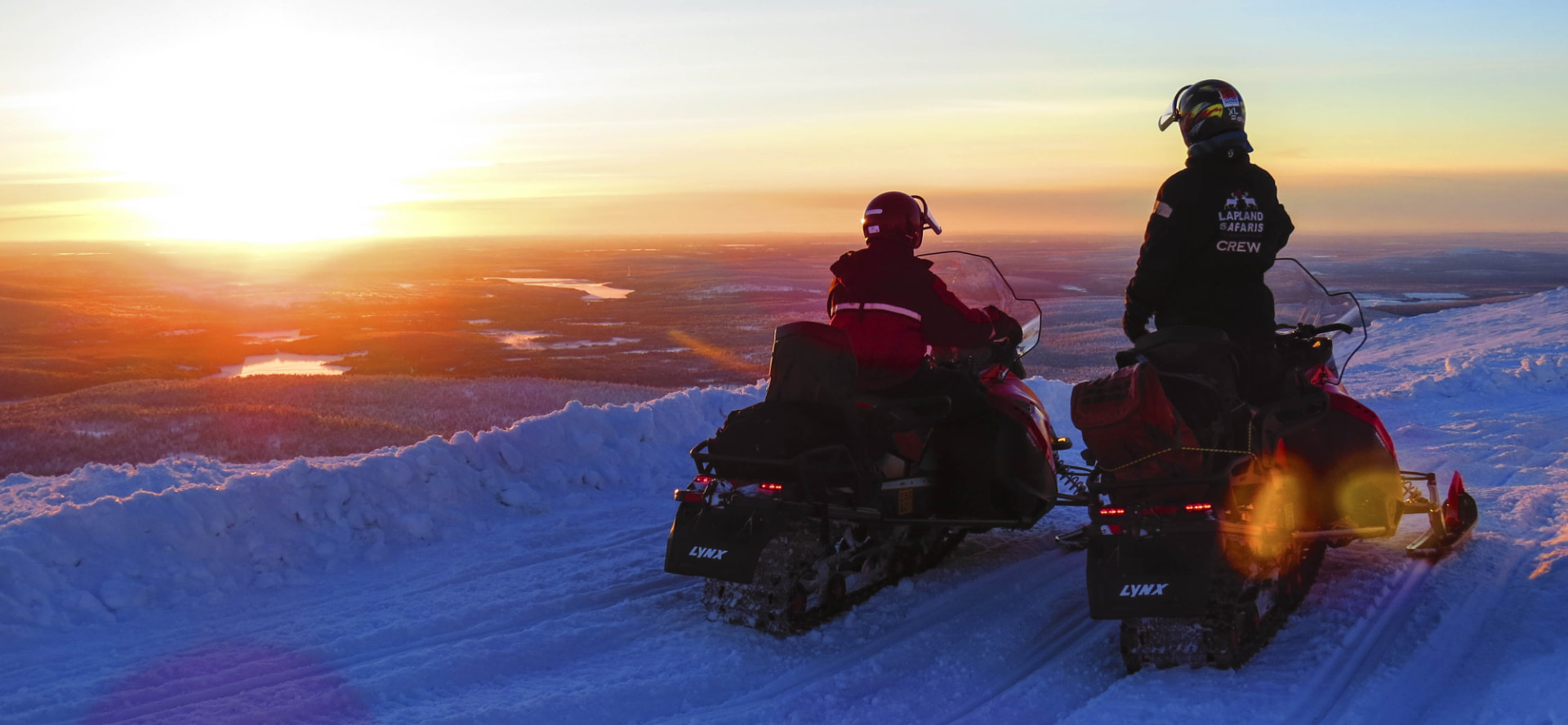 Two guides pausing on snowmobiles at a fell summit above a low winter sun