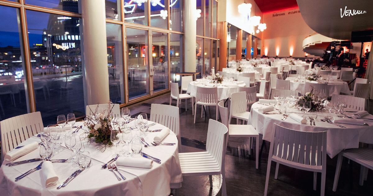 Glass-walled Helsinki dining hall at night with white tables facing the city lights