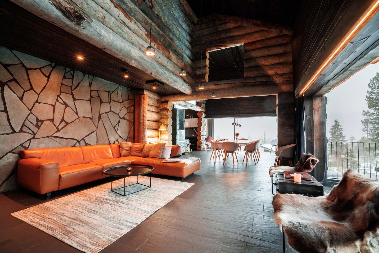 The open living room of a private fell villa with log walls, stone fireplace wall and a long dining table beyond
