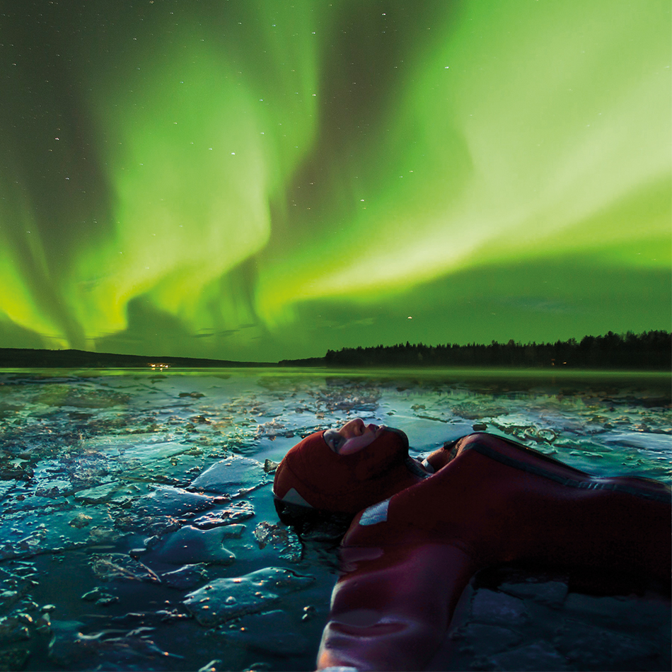 Guest in a survival suit floating among broken lake ice beneath a vivid green aurora