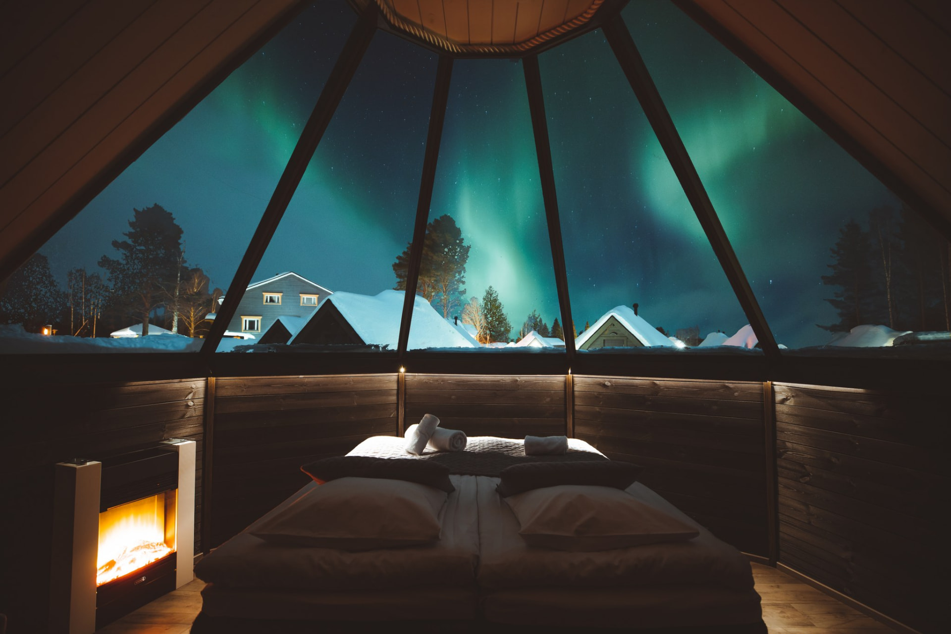 Inside an Apukka aurora cabin: bed and fireplace beneath a glass roof with auroras